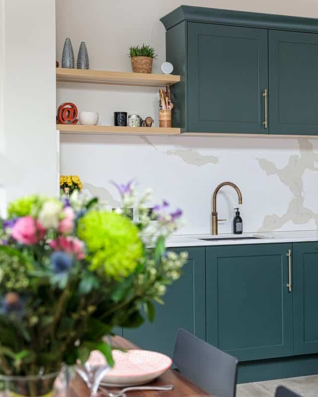 A modern kitchen featuring stylish dark green cabinetry, a sleek brass tap, and an elegant countertop. In the background, there are decorative items on open shelves and a bouquet of colourful flowers in the foreground, adding a touch of freshness to the space. An Arch KBB team member is present, contributing to the design ambiance.