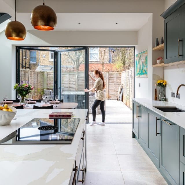 Modern kitchen with open bi-fold doors leading to a garden, featuring an Arch KBB team member interacting with the space. The kitchen showcases sleek cabinetry, a stylish island with a hob, and a dining area set for a meal. Natural light floods the room, highlighting the contemporary design and inviting atmosphere.