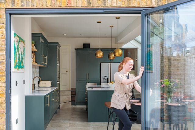 Arch KBB team member interacting with a bi-fold door in a modern kitchen featuring green cabinetry, sleek countertops, and stylish pendant lighting. The open layout enhances natural light and connects the indoor space to an outdoor area with plants.