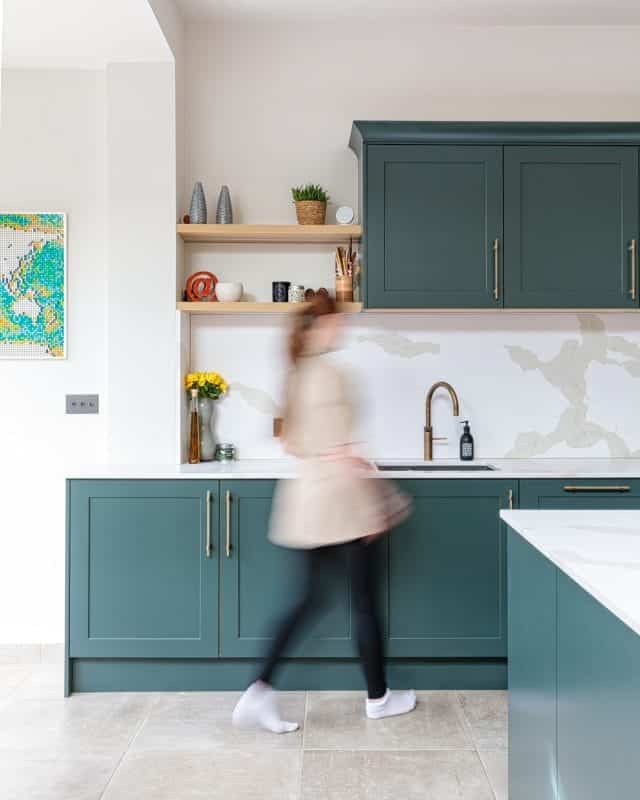 A blurred motion of an Arch KBB team member in a modern kitchen featuring dark green cabinetry, a stylish sink with a gold tap, and open shelving displaying decorative items and plants. The kitchen design showcases a contemporary aesthetic with a light-coloured worktop and a patterned backsplash.