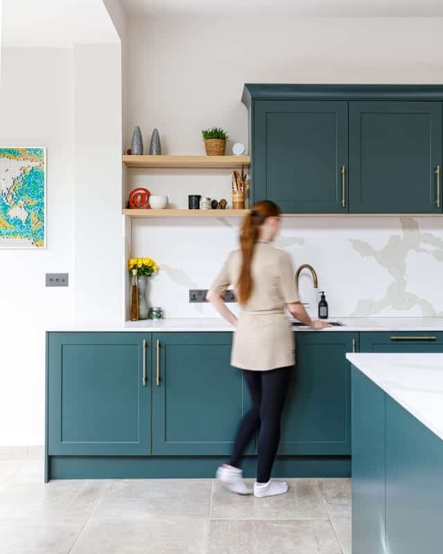 Arch KBB team member standing at a stylish kitchen sink, featuring dark green cabinetry and a light marble backsplash. Decorative items are arranged on open shelves, including a vase with yellow flowers, while the team member is engaged in kitchen tasks.
