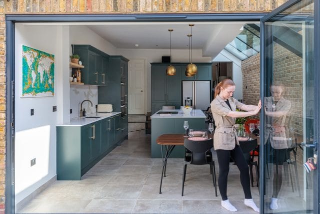 Modern kitchen with green cabinetry and a spacious layout, featuring an Arch KBB team member interacting with the outdoor space. The image showcases a stylish dining area, elegant lighting fixtures, and a large glass door that enhances natural light.
