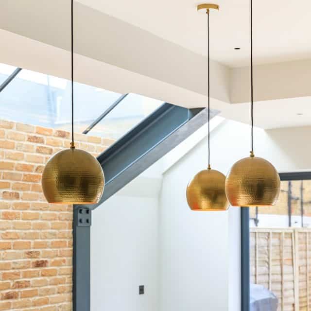 Three modern gold pendant lights hang from the ceiling in a contemporary interior space, featuring a blend of brick and glass elements. The design highlights a stylish and airy atmosphere, showcasing the interplay of light and materials. An Arch KBB team member is present, contributing to the overall aesthetic.