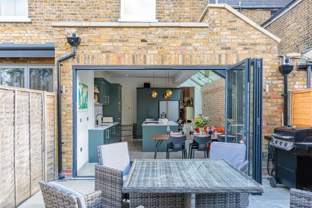 Spacious outdoor dining area featuring a glass-extension kitchen with modern cabinetry and appliances, designed by an Arch KBB team member. The image showcases a seamless transition between indoor and outdoor spaces, with a stylish table setting and contemporary furnishings.