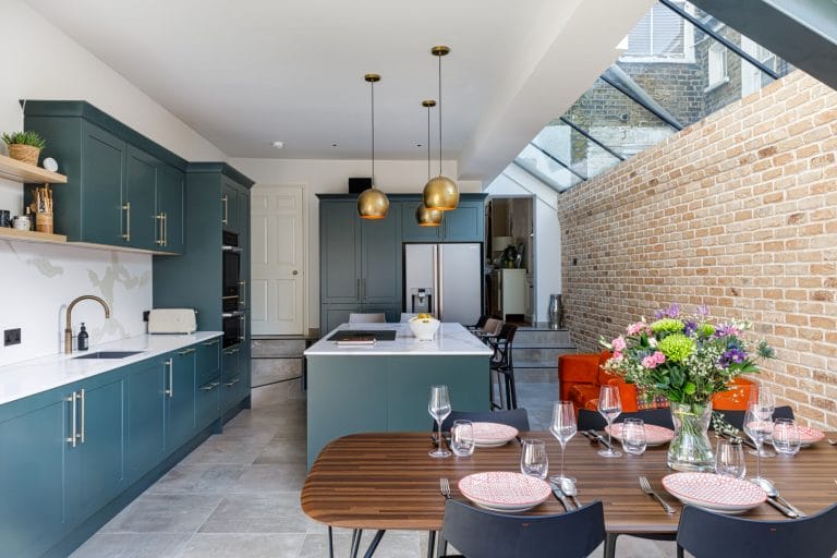 Modern kitchen interior featuring a stylish design with teal cabinetry, marble countertops, and gold pendant lights. A dining table is set with elegant tableware, complemented by a vibrant flower arrangement. The space includes a large window and exposed brick wall, enhancing natural light and creating an inviting atmosphere. An Arch KBB team member is present, showcasing the kitchen's functionality.