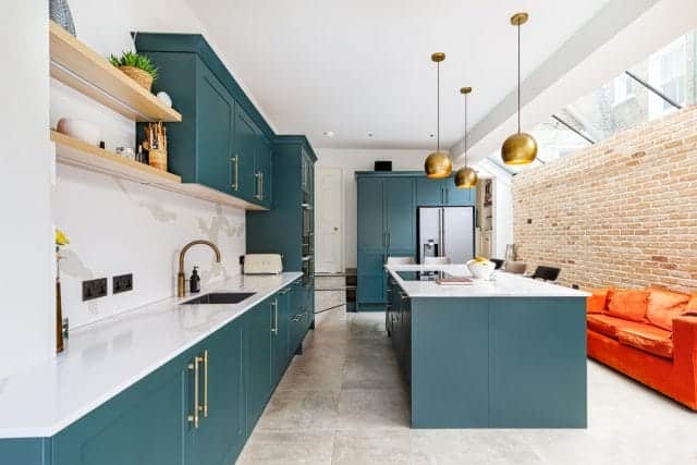 Modern kitchen featuring teal cabinetry, white countertops, and brass pendant lights. The space includes a central island with seating, a brick wall, and an Arch KBB team member engaged in the design process. Natural light floods the room, enhancing the contemporary aesthetic.