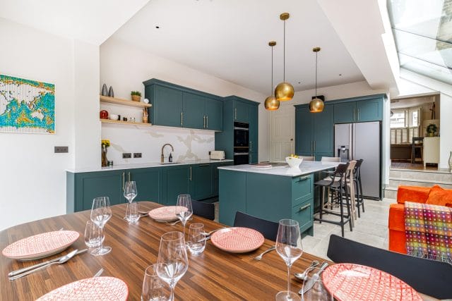 Modern kitchen design featuring teal cabinetry, a spacious island with seating, and a dining table set for six. The room is well-lit with pendant lights and a skylight, showcasing a blend of contemporary style and functionality. An Arch KBB team member is present, enhancing the inviting atmosphere.