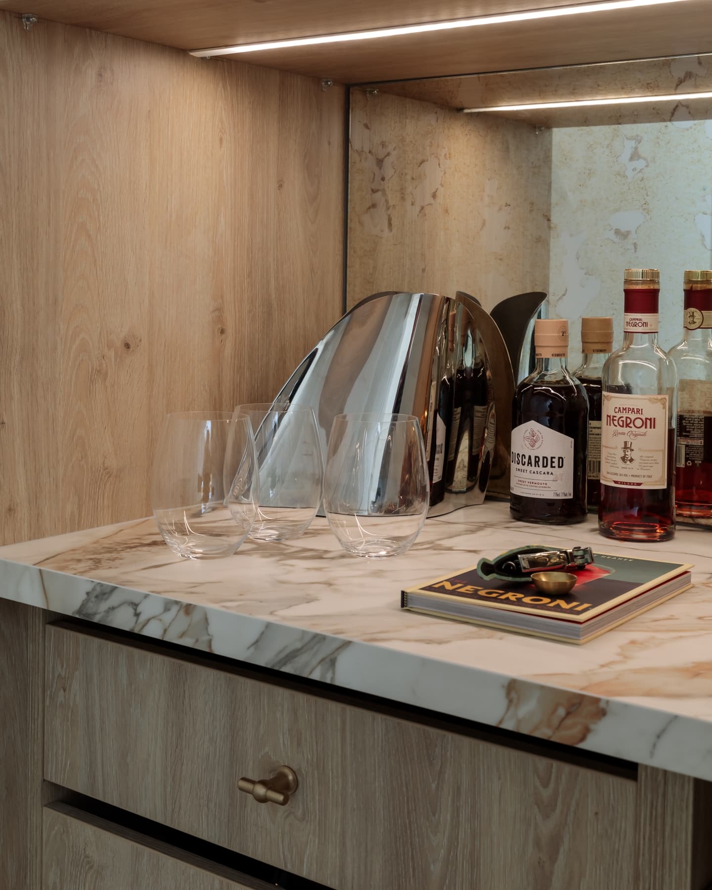 A stylish home bar setup featuring a marble countertop, three clear glasses, and various bottles of spirits including a Negroni recipe book and a cocktail shaker, with an Arch KBB team member present.