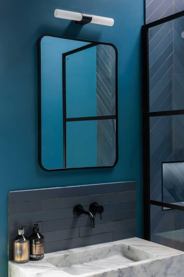 A stylish bathroom featuring a modern black-framed mirror against a blue wall, complemented by sleek taps and geometric tiles. An Arch KBB team member is present, showcasing contemporary design elements and colour coordination.