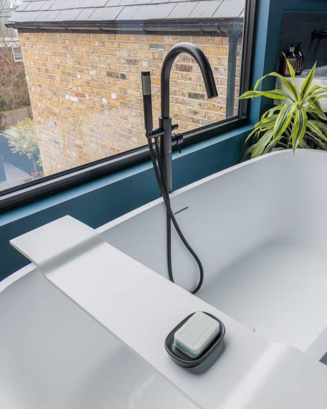 Modern bathroom featuring a sleek freestanding bathtub and a stylish shower fixture. The image showcases natural light streaming through large windows, highlighting the minimalist design elements and greenery in the space. An Arch KBB team member is present, demonstrating attention to detail in a contemporary setting.