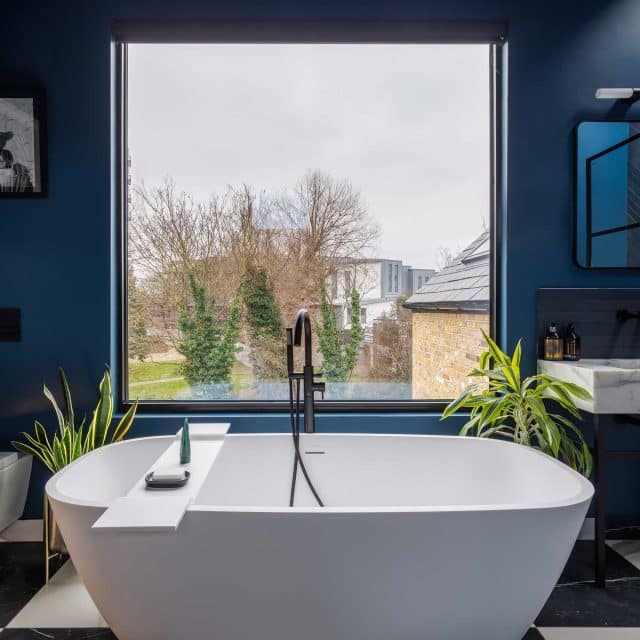 Modern bathroom featuring a freestanding bathtub, framed by large windows offering a view of greenery, with stylish plants and contemporary fixtures. An Arch KBB team member is present, enhancing the design ambiance.