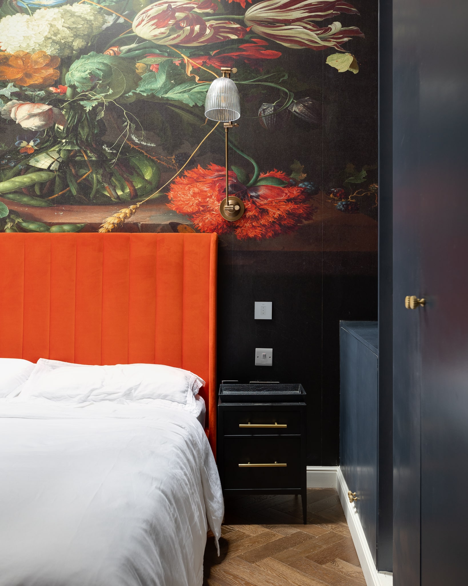 Interior view of a stylish bedroom featuring an orange velvet headboard, crisp white bedding, and a black bedside table. The wall behind is adorned with a vibrant floral mural, complemented by a modern wall light. An Arch KBB team member is present, showcasing the contemporary design elements.