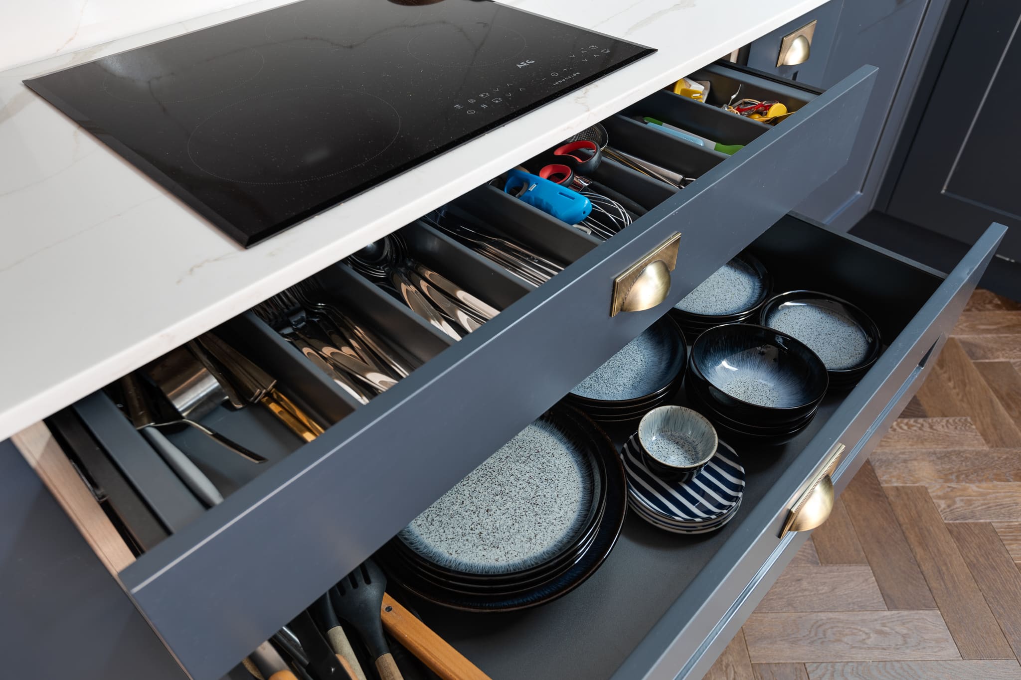 Interior view of a modern kitchen featuring a well-organised drawer filled with cutlery and kitchen utensils, alongside another drawer displaying an assortment of stylish plates and bowls. An induction cooktop is visible on the countertop above. Arch KBB team member's workspace highlighted.
