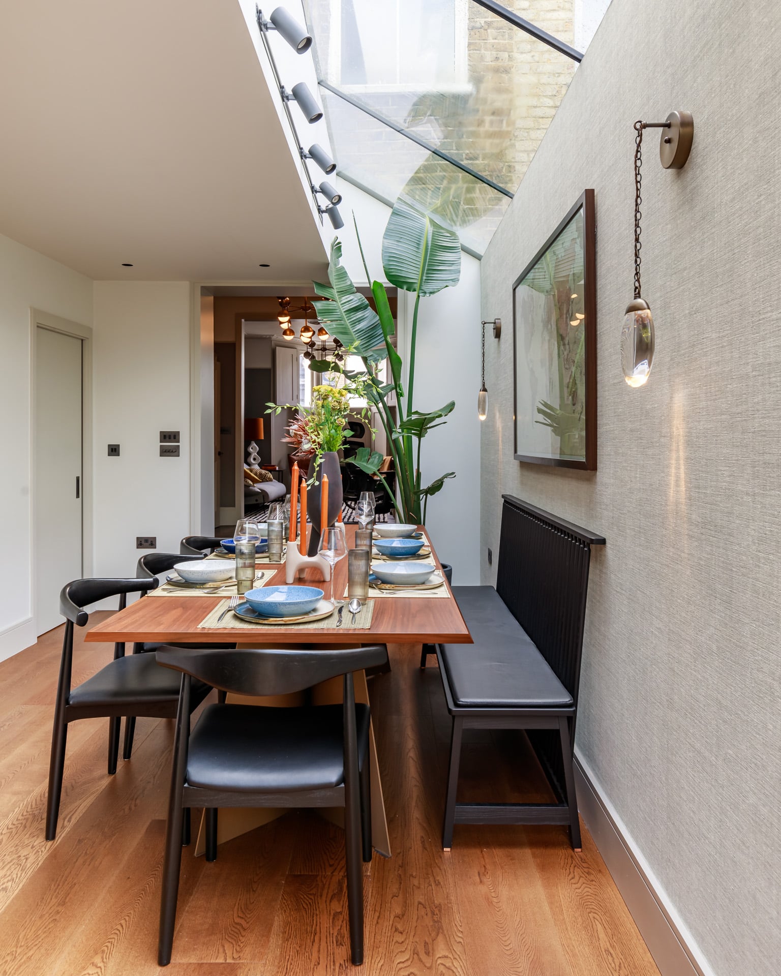 Modern dining area featuring a wooden table set for a meal, complete with blue plates, glassware, and decorative elements. Lush green plants add a touch of nature, while stylish lighting fixtures and wall art enhance the contemporary design. An Arch KBB team member can be seen in the background, contributing to the inviting atmosphere.