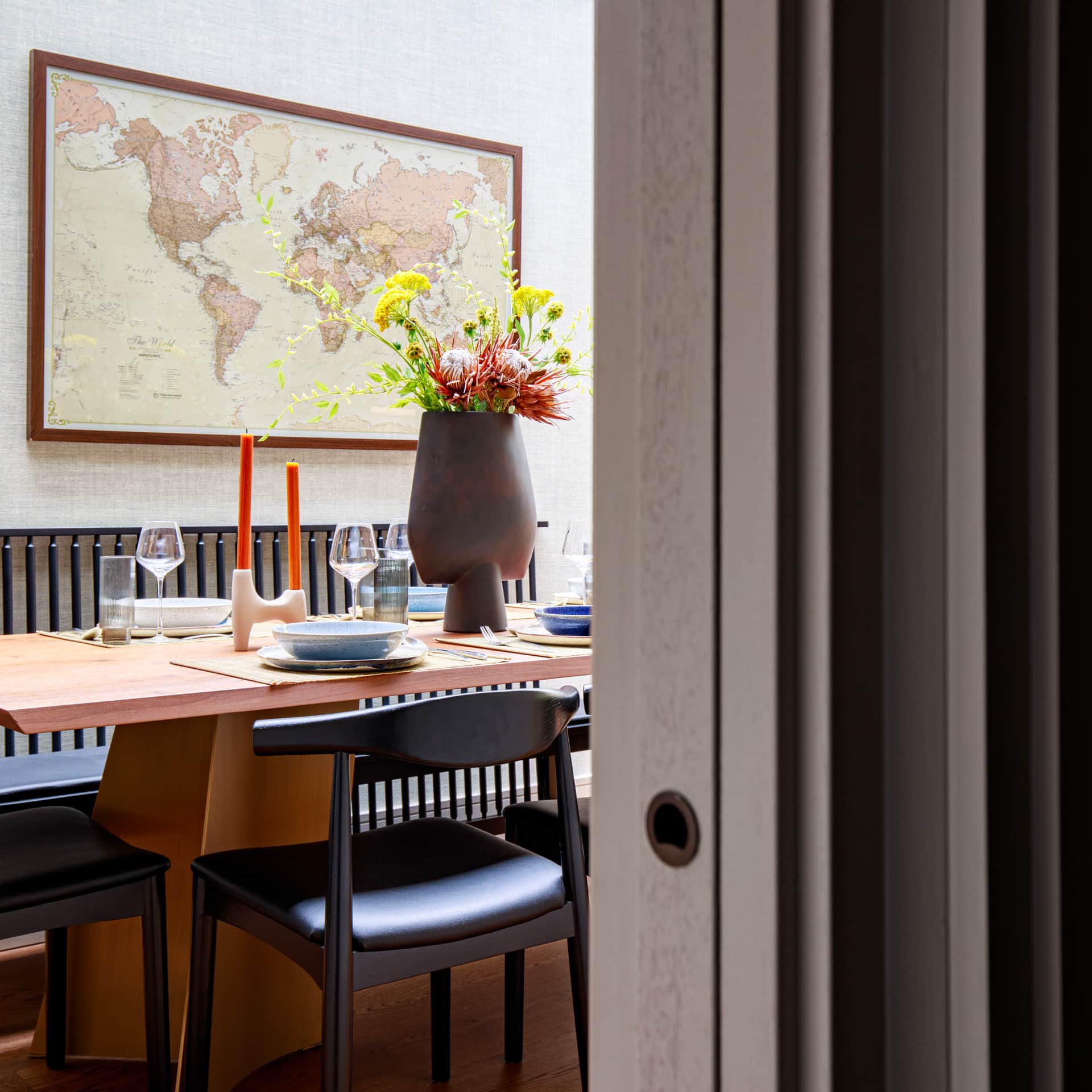 Interior view of a dining area featuring a wooden table set for a meal, with stylish tableware, glasses, and decorative elements including a large vase filled with flowers. A world map is displayed on the wall, enhancing the contemporary decor. An Arch KBB team member is positioned in the scene, contributing to the inviting atmosphere.
