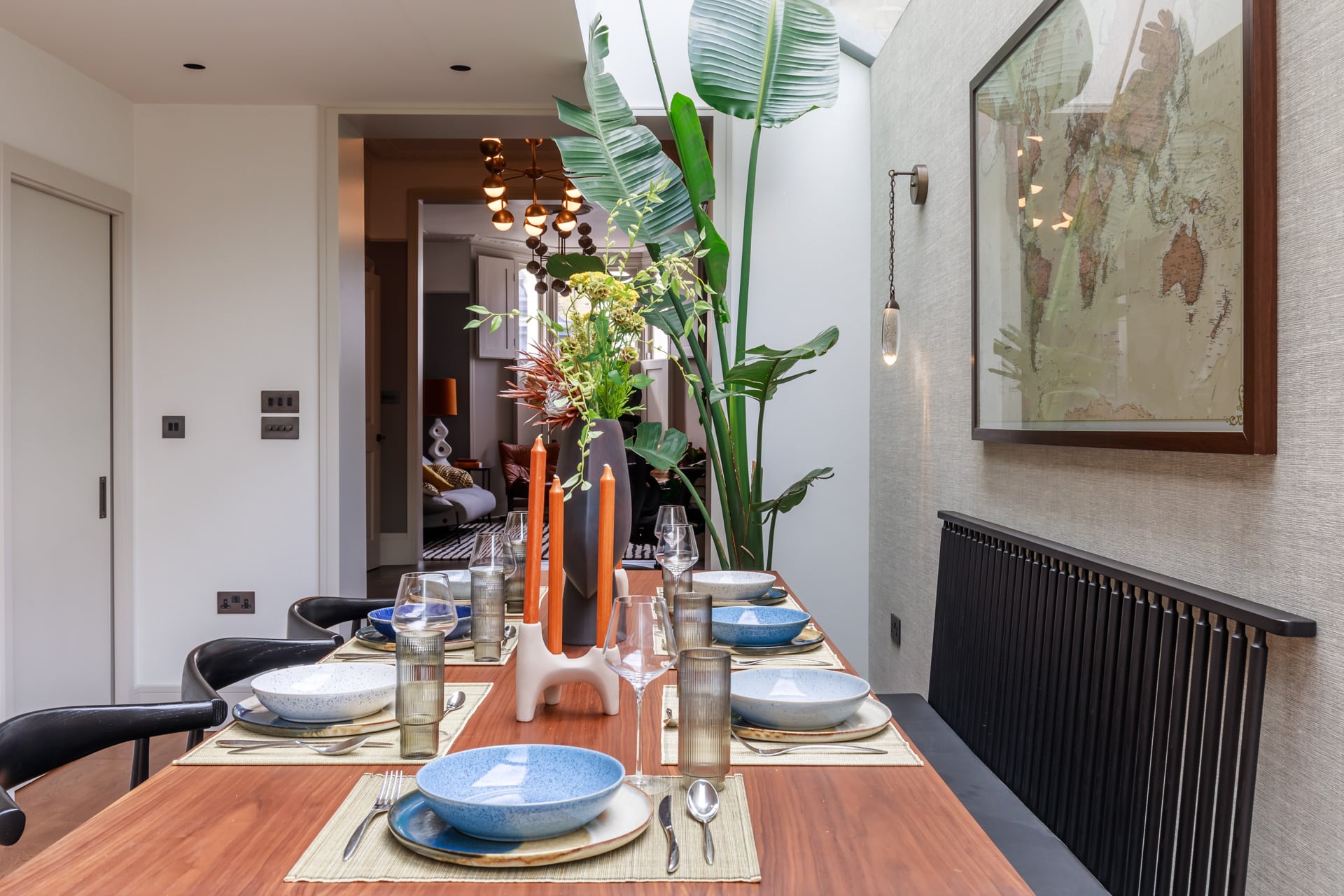 Dining room with a wooden table set for a meal, featuring blue and white crockery, glasses, and decorative candles. A large plant adds greenery to the space, while a piece of artwork adorns the wall. An Arch KBB team member is present, enhancing the inviting atmosphere.