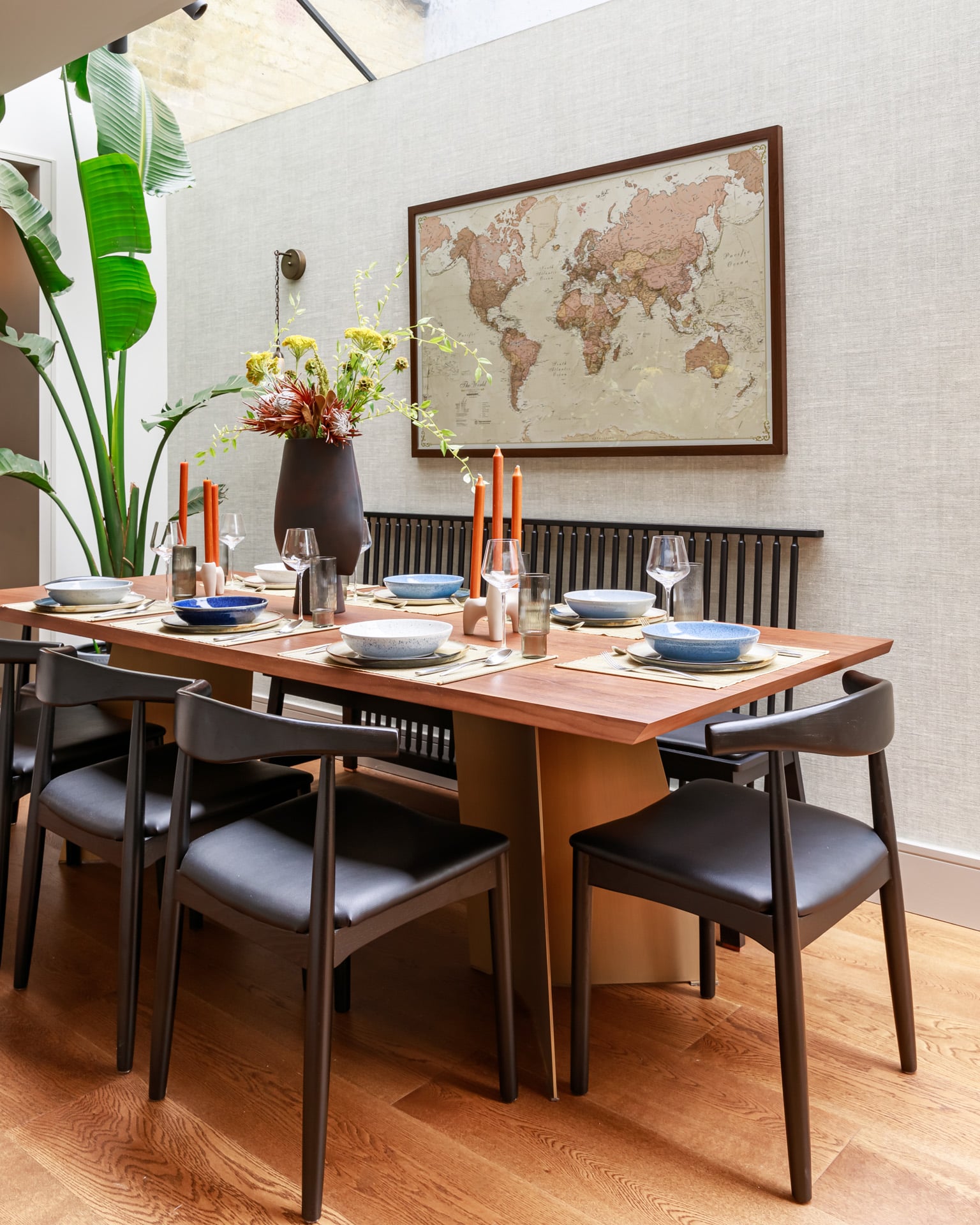 A stylish dining area featuring a wooden table set for a meal with blue and white tableware, glassware, and decorative candles. A large vase with flowers adds a touch of elegance, while a world map hangs on the wall. An Arch KBB team member is present, contributing to the inviting atmosphere. Lush greenery completes the modern decor.