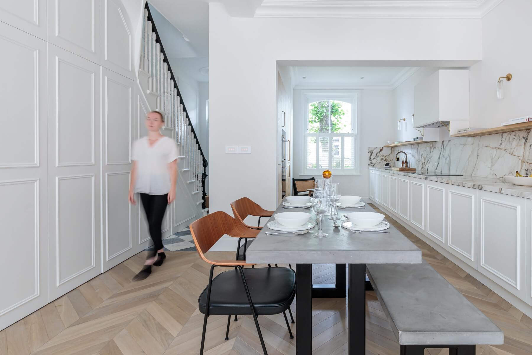 A modern kitchen and dining area featuring a marble-topped table set for a meal, with wooden and black chairs, and a light-filled space. An Arch KBB team member walks past the dining area, showcasing a blend of contemporary design and functionality.