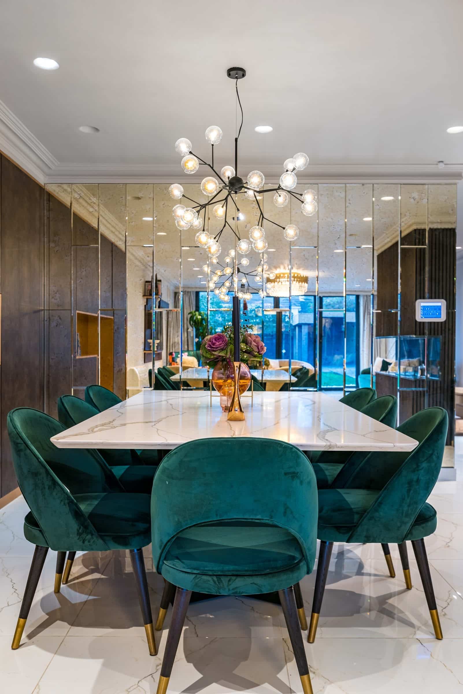 Modern dining area featuring a marble table surrounded by plush green chairs, complemented by an elegant chandelier and decorative floral arrangement. The room is illuminated by natural light from large windows, with mirrored walls adding depth to the space. An Arch KBB team member can be seen in the background, contributing to the inviting atmosphere.