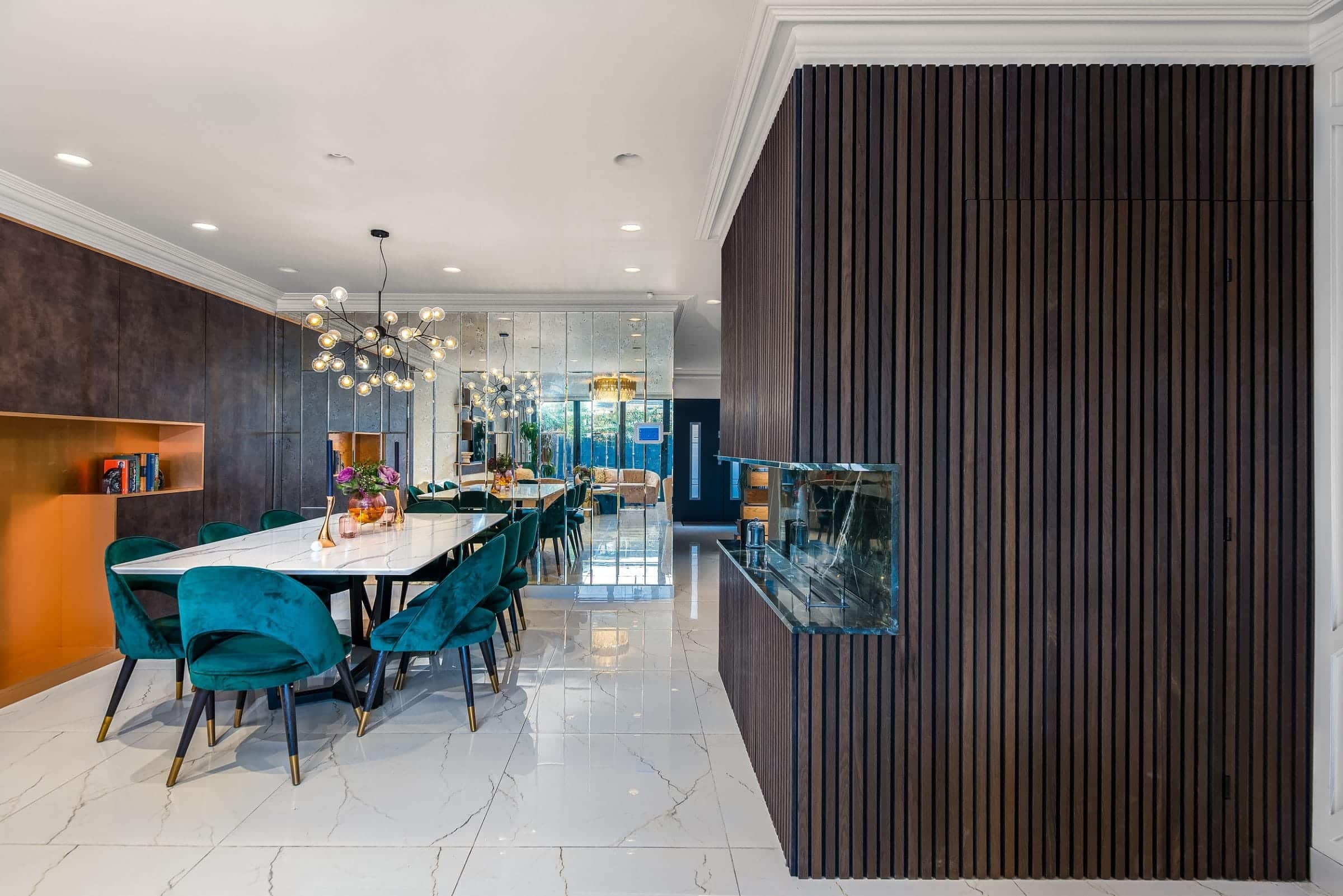 Modern dining area featuring a large white table surrounded by teal chairs, complemented by a stylish chandelier. The room showcases a mirrored wall that reflects the vibrant decor and a sleek wooden feature wall. An Arch KBB team member is present, enhancing the inviting atmosphere of the space.