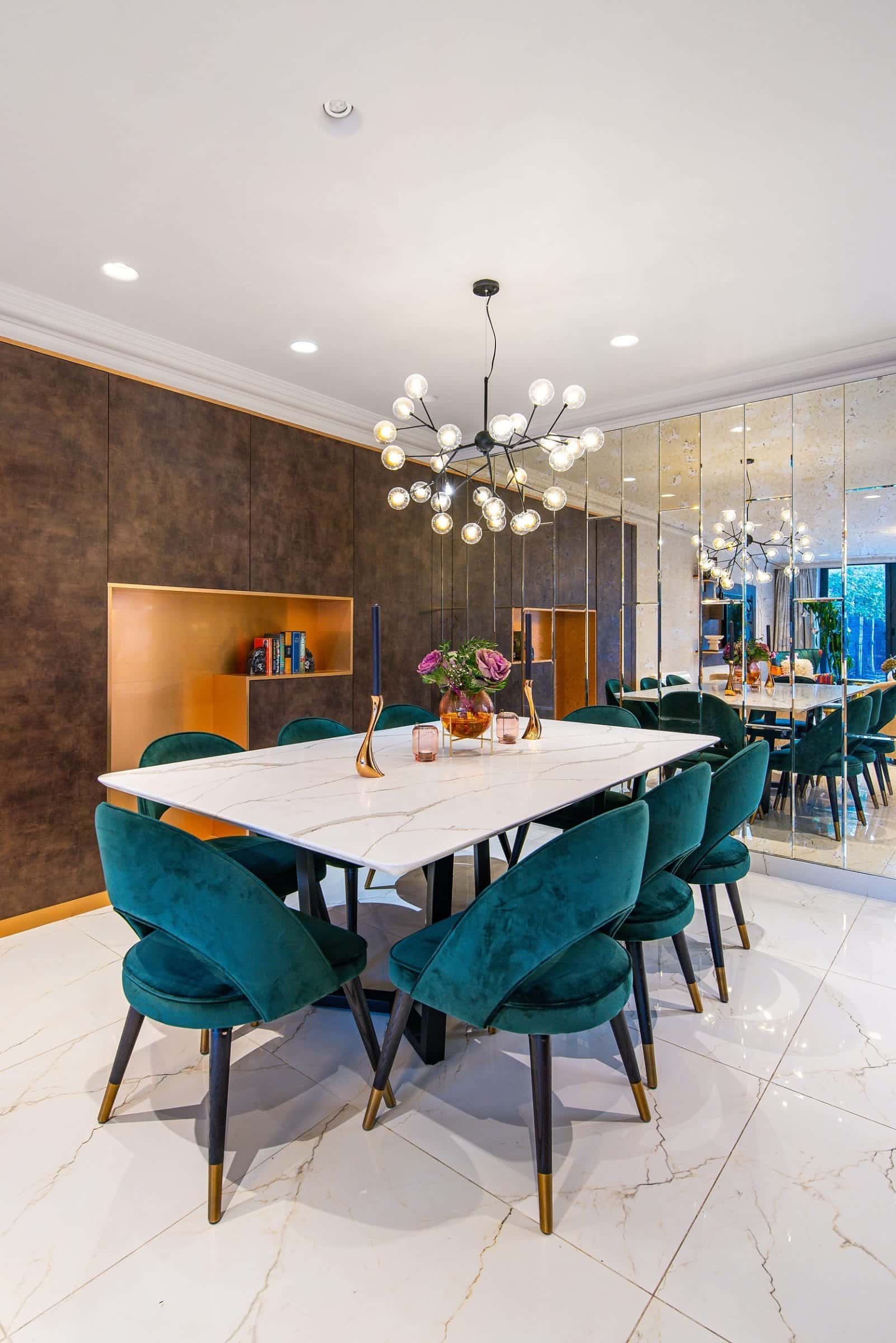 A stylish dining area featuring a marble table surrounded by dark green upholstered chairs, accented by a contemporary chandelier. The wall showcases a mix of reflective mirrors and a warm-toned feature panel with shelves. An Arch KBB team member is present, adding a touch of professionalism to the setting.