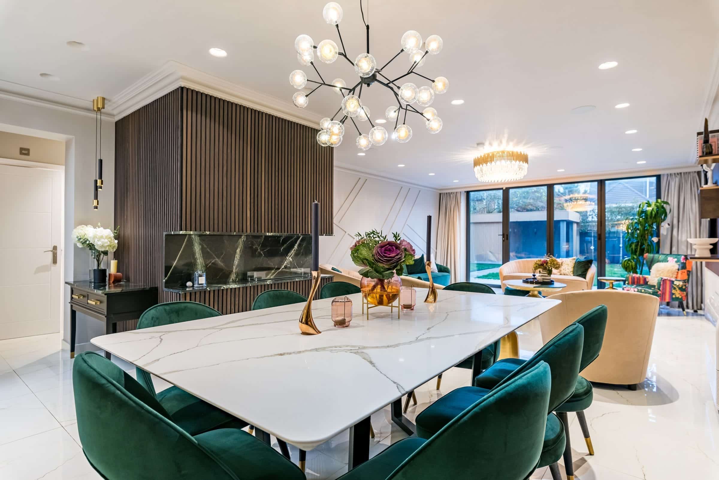 Modern dining area featuring a sleek marble table surrounded by plush green chairs. The room is illuminated by stylish chandeliers, with a contemporary fireplace and large windows allowing natural light to flood in. An Arch KBB team member is present, enhancing the inviting atmosphere of the space.