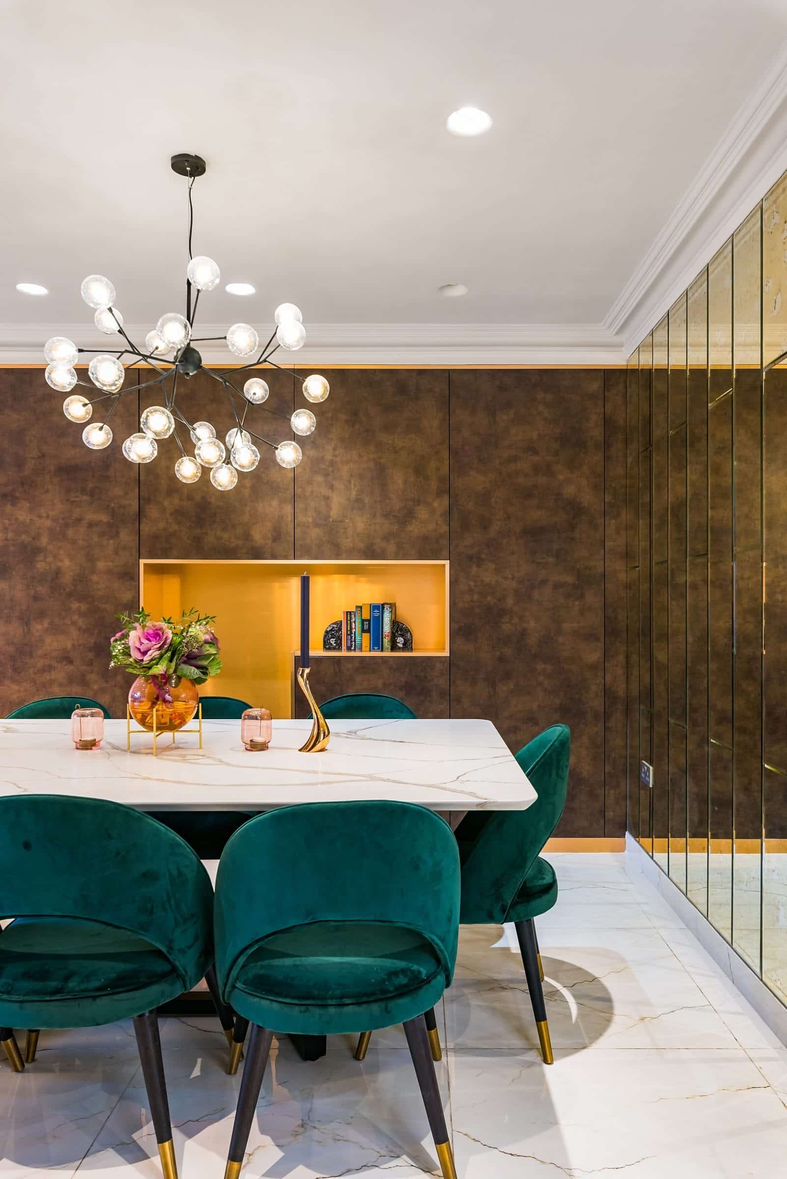 Modern dining room featuring a marble table surrounded by green velvet chairs, complemented by a unique chandelier. A decorative display with flowers, candles, and books is visible in a stylish wall niche, while mirrored walls add a touch of elegance. An Arch KBB team member is present, contributing to the design ambiance.