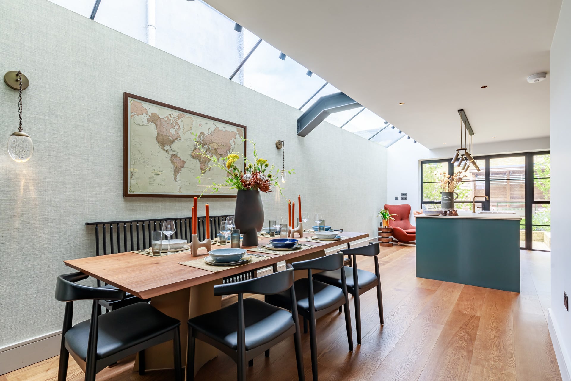 Stylish dining area featuring a wooden table set with tableware and floral arrangements, complemented by a world map on the wall. An Arch KBB team member is present, showcasing a modern kitchen with a green island and natural light streaming through skylights.