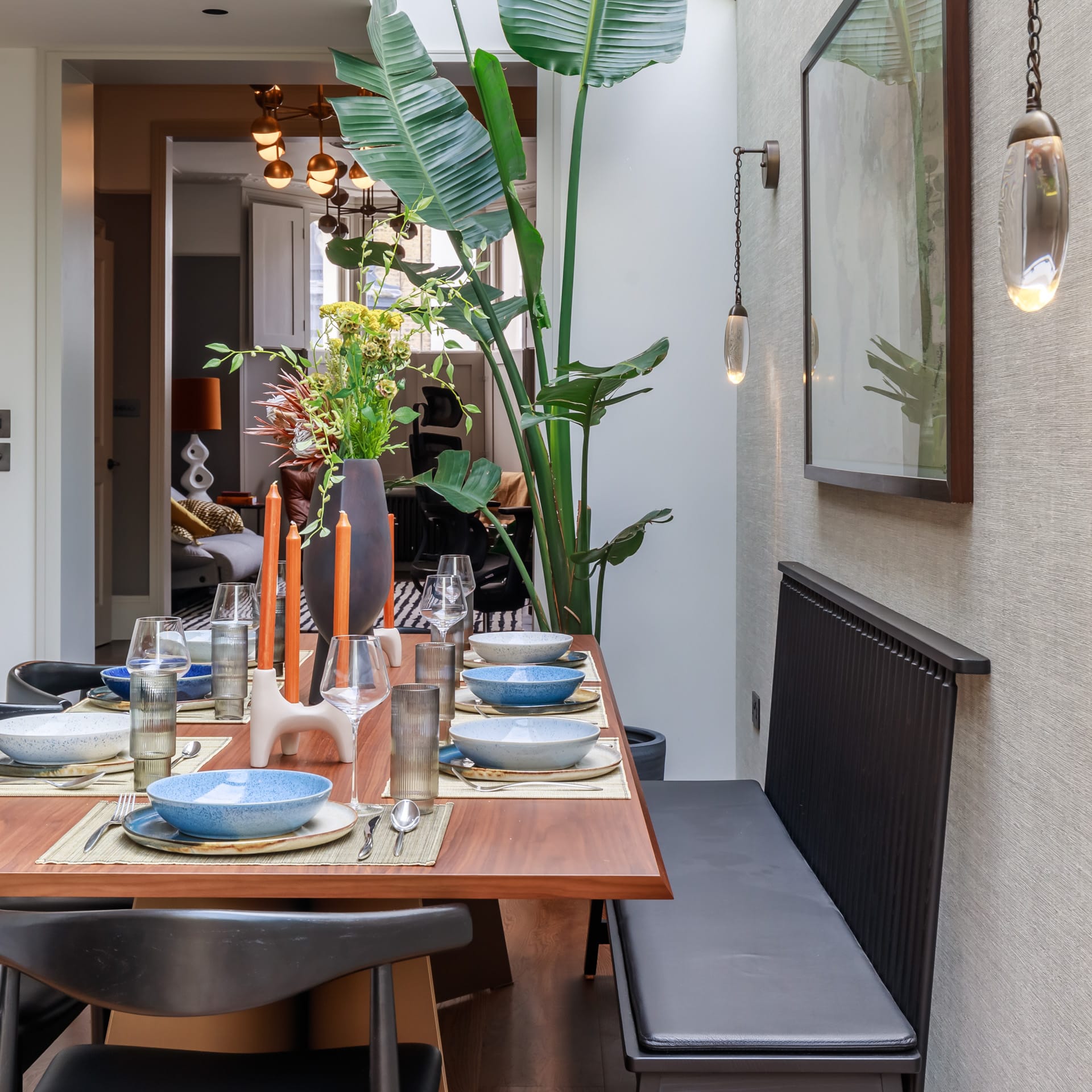A modern dining area featuring a wooden table set for a meal with blue bowls, glasses, and candles. Decorative plants and a stylish wall art piece enhance the ambiance, while an Arch KBB team member is present in the background, contributing to the inviting atmosphere.