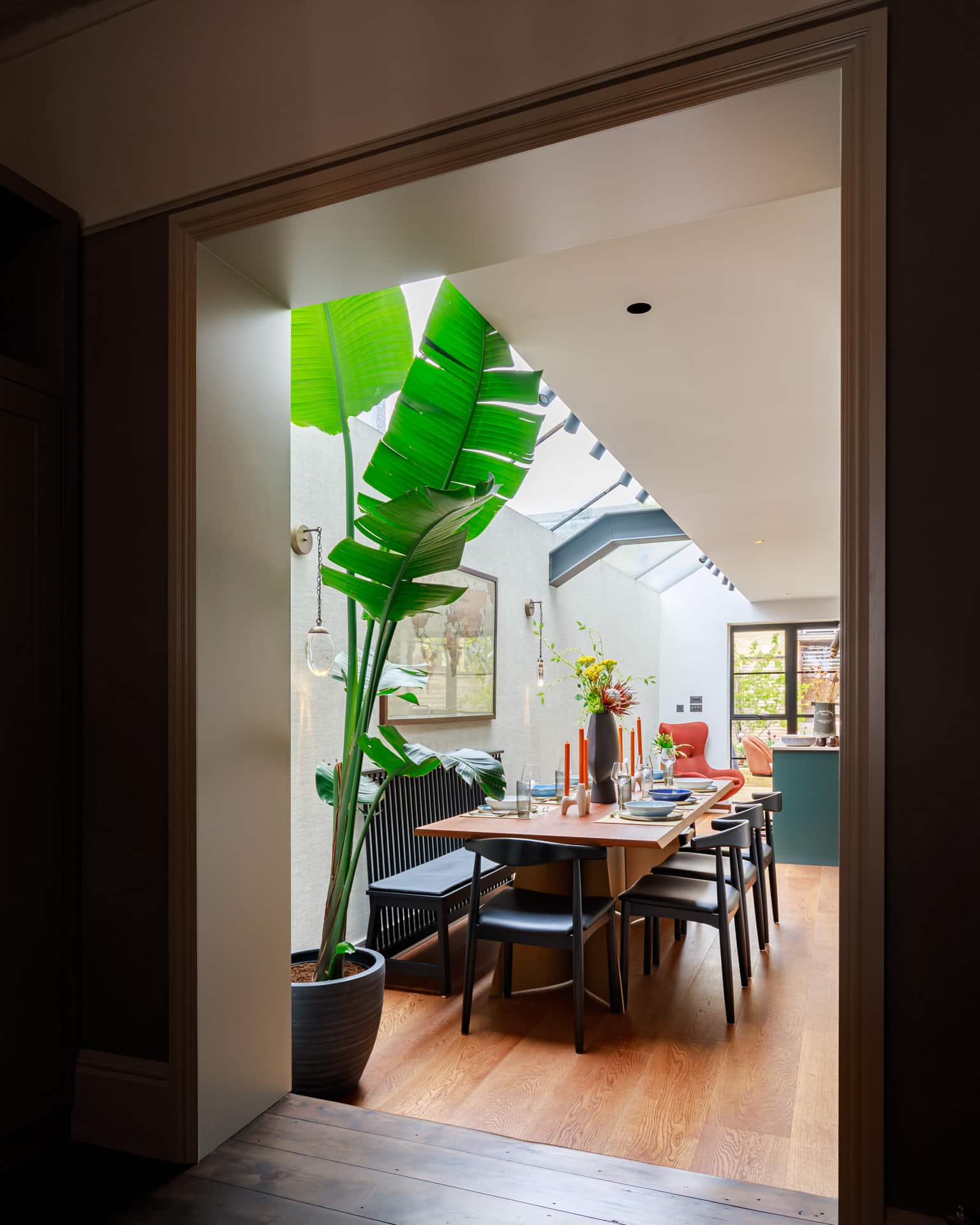 Interior view of a stylish dining area featuring a large table set for a meal, surrounded by black chairs. A tall, vibrant plant adds a touch of greenery, while natural light streams in through a skylight. An Arch KBB team member is present, enhancing the inviting atmosphere of the space.
