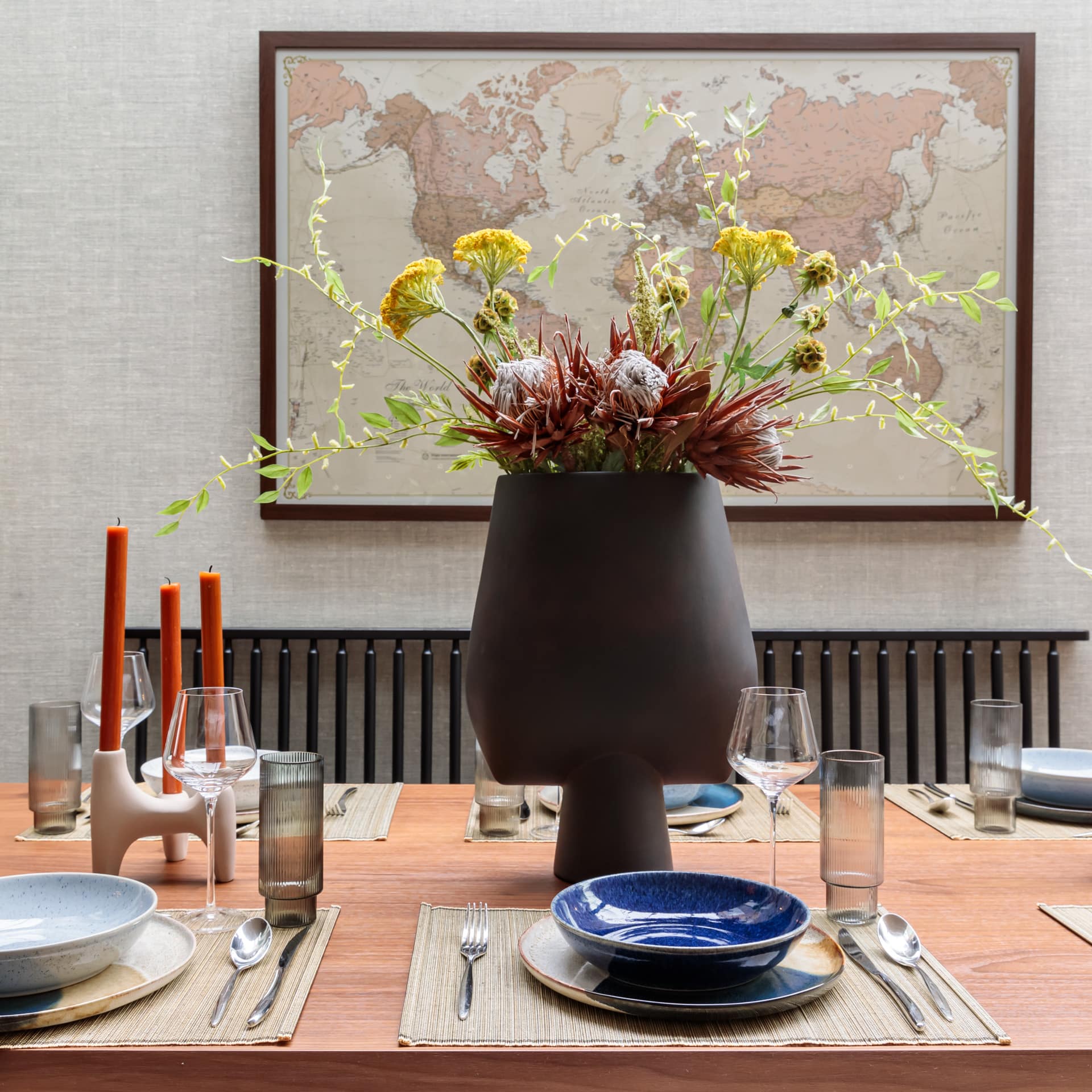 A beautifully set dining table featuring an elegant arrangement of dried flowers in a large vase, surrounded by glassware and tableware. The backdrop includes a world map, enhancing the sophisticated atmosphere of the space. An Arch KBB team member is present, contributing to the inviting setting.