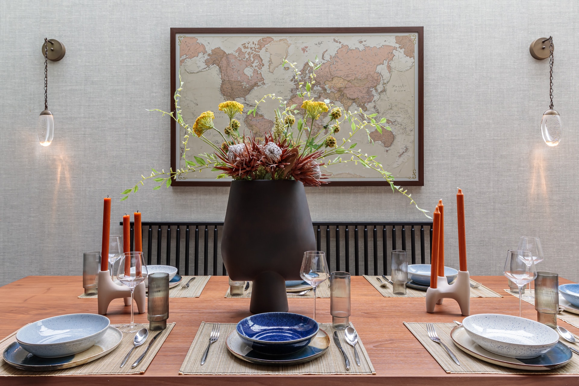 A stylish dining table set for a meal, featuring a large vase with mixed dried flowers at the centre. The table is adorned with various plates, glasses, and cutlery, complemented by orange candles. A world map hangs on the wall behind, adding a touch of sophistication to the space. An Arch KBB team member is present, contributing to the inviting atmosphere.