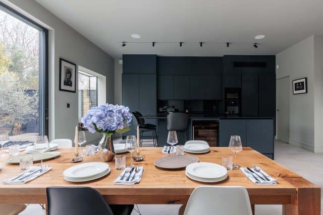 Modern dining area featuring a wooden table set with plates and glassware, complemented by a vase of blue hydrangeas. The stylish kitchen in the background showcases dark cabinetry and contemporary appliances. An Arch KBB team member is seated at the counter, enhancing the inviting atmosphere.