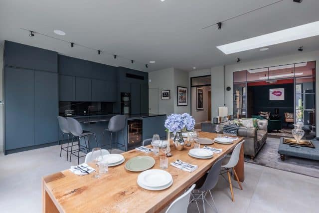 Modern open-plan kitchen and dining area featuring a wooden dining table set for a meal, with elegant tableware and a vase of flowers. The kitchen showcases contemporary cabinetry in dark tones, with sleek appliances and stylish bar stools. An Arch KBB team member is present, enhancing the inviting atmosphere of the space.