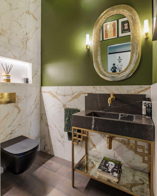Modern bathroom design featuring elegant marble tiles, a stylish black toilet, and a sophisticated washbasin with a gold faucet. The walls are painted in a rich green hue, complemented by an ornate mirror and ambient lighting. An Arch KBB team member is present, showcasing the contemporary decor and design elements.