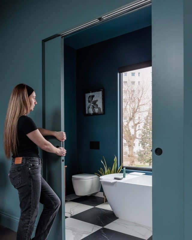 Arch KBB team member opening a sliding door in a modern bathroom with dark blue walls, featuring a freestanding bathtub, a toilet, and a window with a view of trees outside.