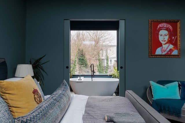 A stylish bedroom featuring a comfortable bed with decorative cushions, a modern freestanding bathtub visible through a large window, and a vibrant artwork on the wall. An Arch KBB team member is present, showcasing the interior's design elements and tranquil atmosphere.