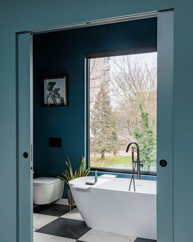 Modern bathroom design featuring a freestanding bathtub, sleek black tap, and a large window with a view of greenery. An Arch KBB team member is present, showcasing the stylish interior with a contemporary colour palette and framed artwork on the wall.