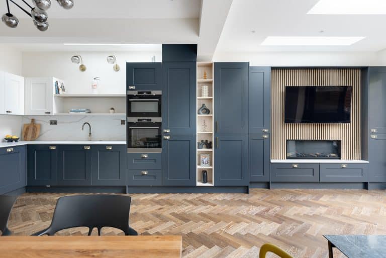 Modern kitchen design featuring navy blue cabinetry, integrated appliances, and a stylish wooden floor. The space includes a sleek oven, open shelving, and a wall-mounted television, creating a contemporary and functional environment. An Arch KBB team member is present, contributing to the design process.