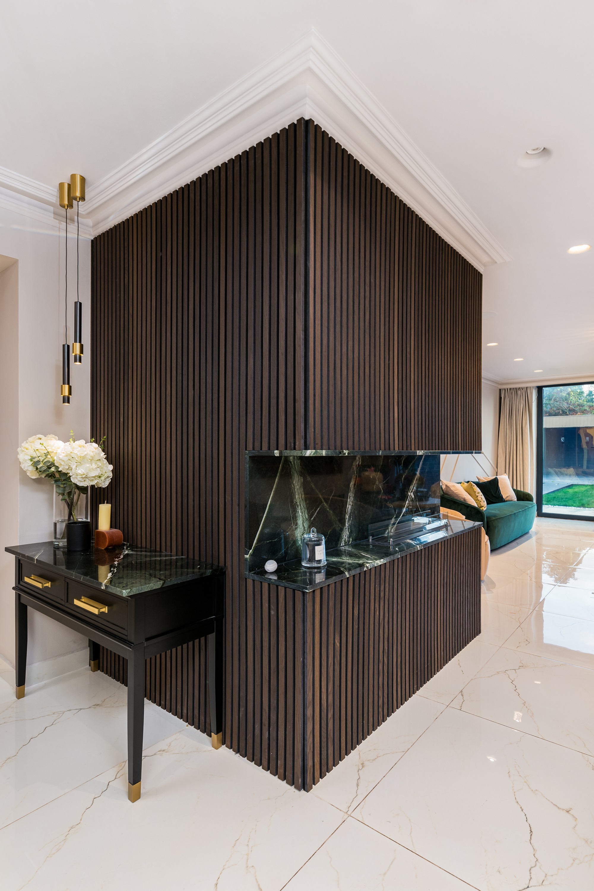 Interior view of a modern living space featuring a wooden feature wall with vertical slats, a sleek black console table adorned with decorative items, and a stylish green sofa. Natural light flows in through large windows, enhancing the elegant atmosphere. An Arch KBB team member is present, contributing to the design aesthetic.