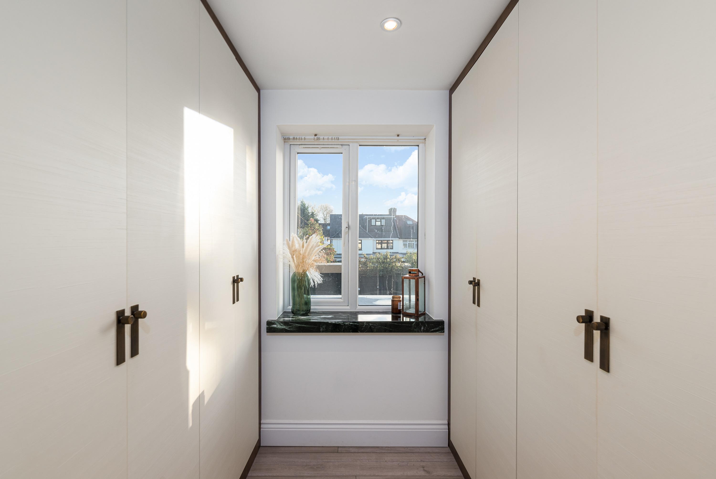 Interior view of a modern hallway featuring built-in wardrobes with sleek handles, a window allowing natural light, and a decorative arrangement of dried flowers on a dark countertop. An Arch KBB team member is present, showcasing the stylish design and functionality of the space.