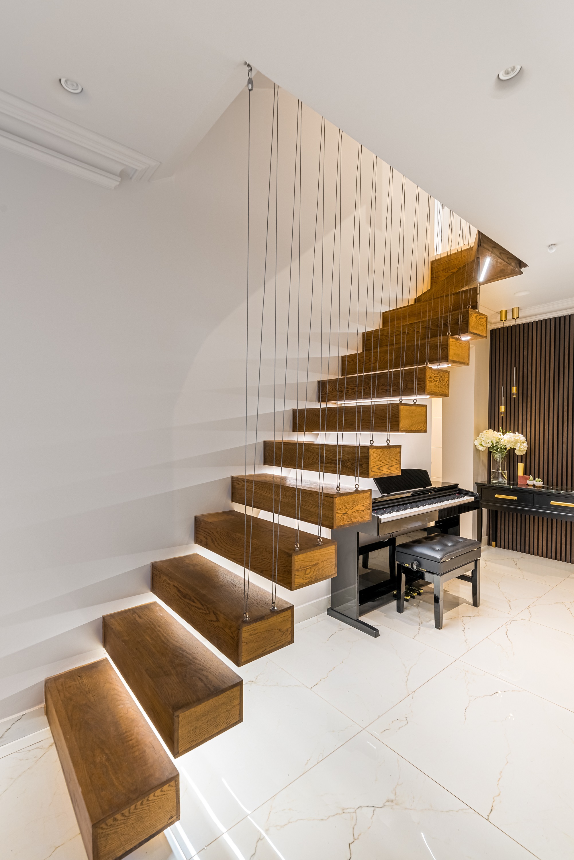 Stylish interior featuring a modern suspended staircase with wooden steps, accompanied by a piano and elegant decor. An Arch KBB team member is present, enhancing the inviting atmosphere of the space.