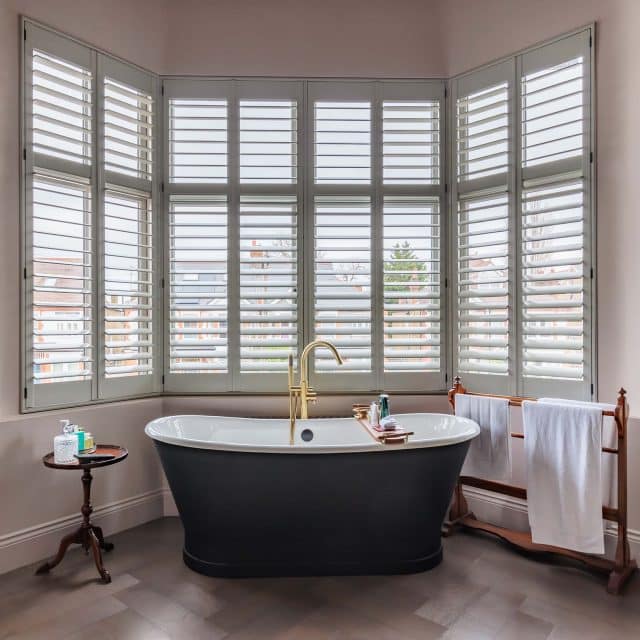 Elegant bathroom featuring a freestanding bathtub with a gold faucet, surrounded by stylish window shutters. A small side table holds toiletries, while a wooden towel rack displays neatly hung towels. The design combines modern and classic elements, creating a serene atmosphere. An Arch KBB team member is present to enhance the scene.