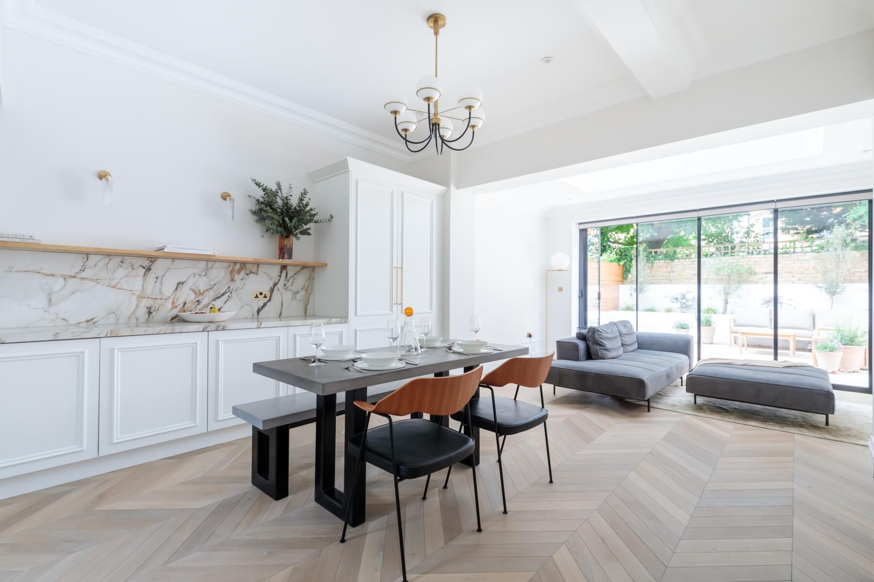 Modern dining area featuring a stylish table set for four, surrounded by elegant chairs. The room boasts a light colour palette with wooden flooring and a chic chandelier. A contemporary kitchen area with marble accents is visible, along with large glass doors leading to an outdoor space. An Arch KBB team member is present, showcasing the inviting atmosphere of the home.