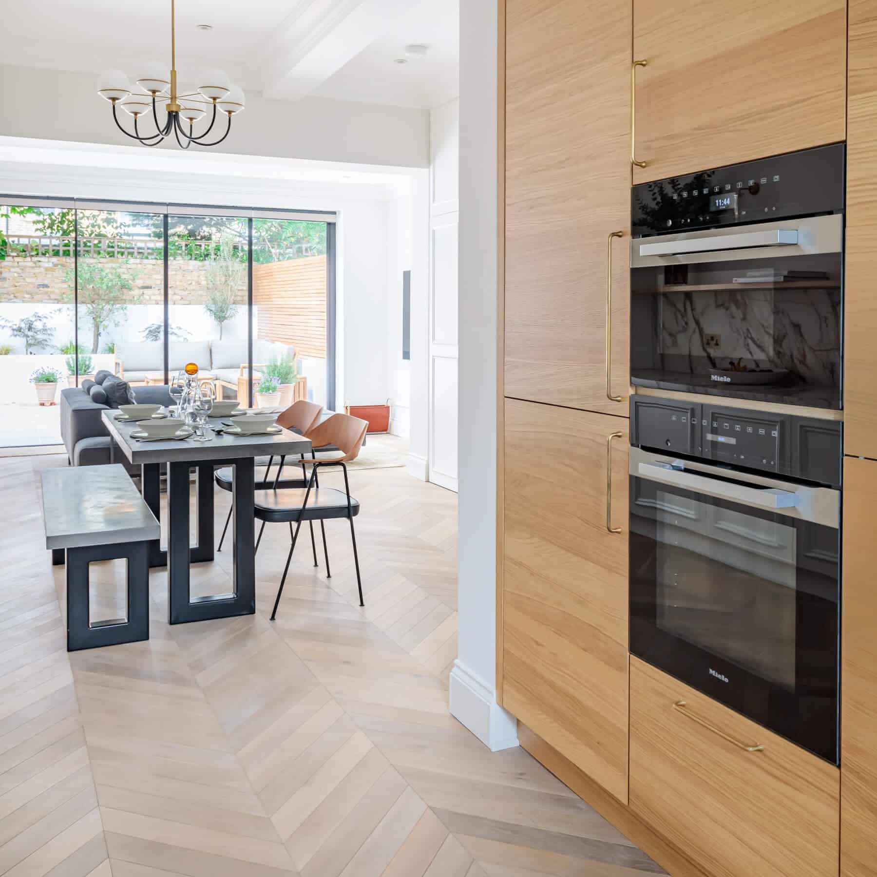 Modern kitchen with integrated appliances featuring a sleek design, showcasing a dining area with a wooden table set for a meal. An Arch KBB team member is present, enhancing the inviting atmosphere of the space filled with natural light and stylish decor.