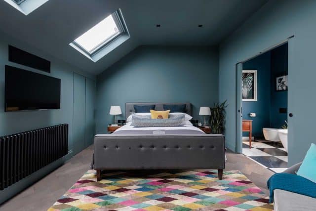 Modern bedroom design featuring a grey upholstered bed with decorative pillows, surrounded by teal walls and natural light from a skylight. The space includes stylish bedside lamps, a vibrant patterned rug, and a glimpse of an adjoining bathroom. An Arch KBB team member is present, showcasing the contemporary interior decor.