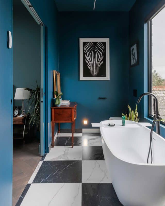 Modern bathroom featuring a freestanding bathtub, stylish black and white tiled flooring, and a deep blue accent wall. The space includes a wooden cabinet, decorative plants, and artwork, creating a sophisticated atmosphere. An Arch KBB team member is present, enhancing the design experience.