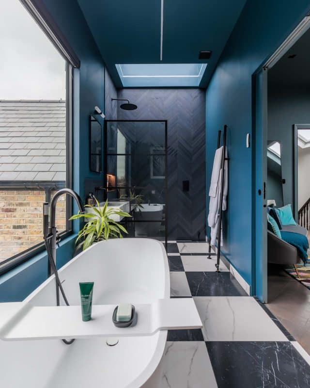 Modern bathroom design featuring a freestanding bathtub, stylish black and white tiled floor, and blue walls. An Arch KBB team member is present, and large windows provide natural light. The space includes a glass shower enclosure and contemporary fixtures, creating a sleek and inviting atmosphere.