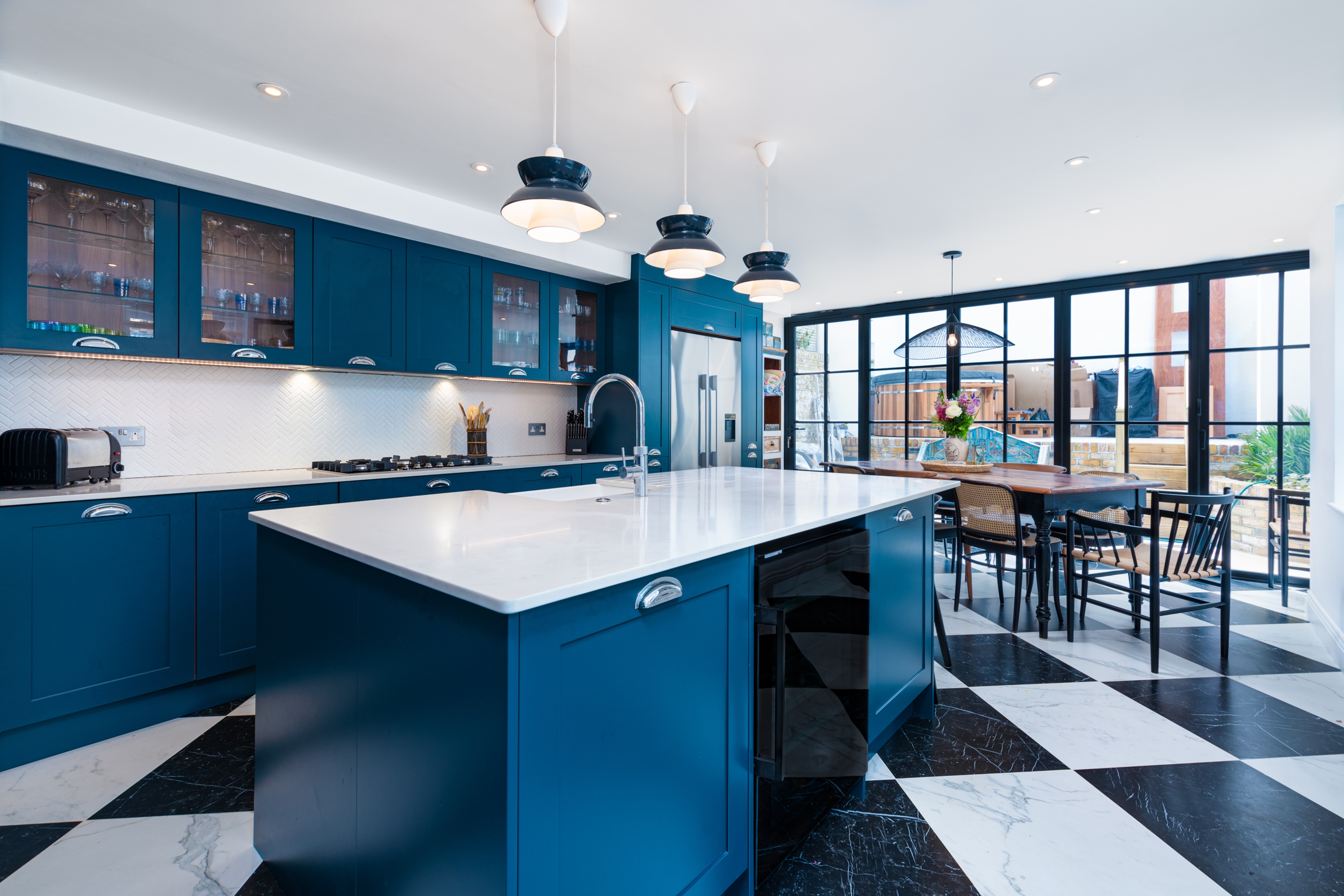 Modern kitchen featuring a stylish blue cabinetry design, a large white island with a sink, and black and white tiled flooring. The space is illuminated by pendant lights, and there is a dining area with wooden chairs visible through large glass doors. An Arch KBB team member is present, adding a professional touch to the scene.