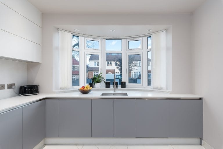 Modern kitchen featuring a sleek design with grey cabinetry, a white countertop, and a large bay window allowing natural light. The space includes a bowl of fruit and a plant, enhancing the inviting atmosphere. An Arch KBB team member is present, showcasing a stylish and functional kitchen environment.
